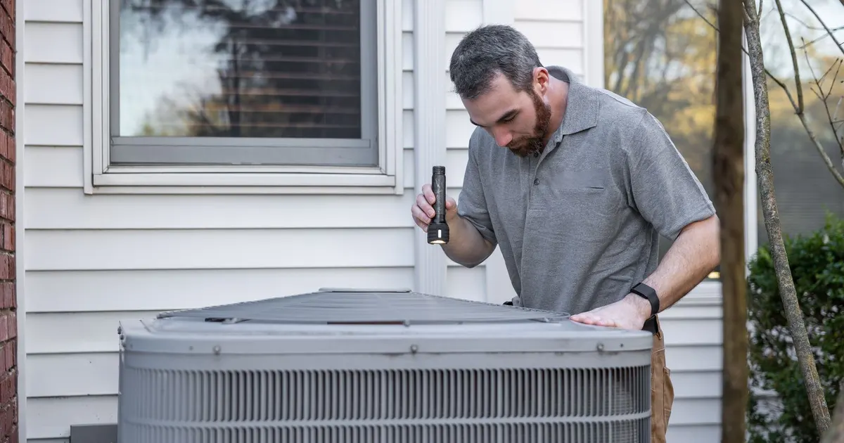 HVAC technician inspecting an outdoor air conditioning condenser unit with a flashlight